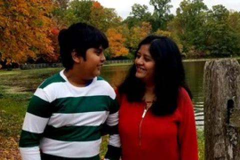 印度遗产的少年，穿着绿色和白色条纹衬衫，看着他的母亲站在五颜六色的秋天树前的红色衬衫上