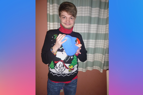 一个年轻人站在一个黑色的毛衣和圣诞树的图形与史努比和伍德斯托克音乐节。与一个红色的衬衫。他对着镜头微笑,手里拿着一个玩具收音机闹钟