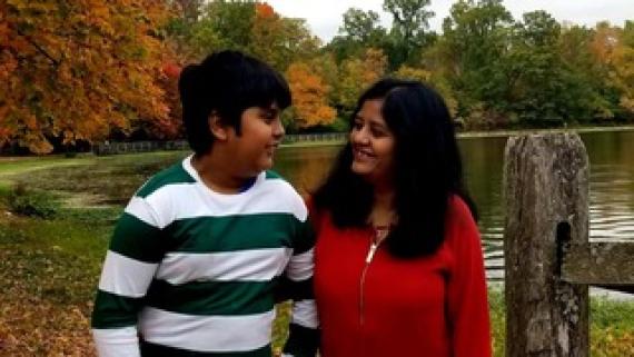 印度遗产的少年，穿着绿色和白色条纹衬衫，看着他的母亲站在五颜六色的秋天树前的红色衬衫上